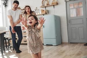 finalmente nos conocimos. una niña juguetona se divierte corriendo en la cocina durante el día frente a su madre y su padre foto