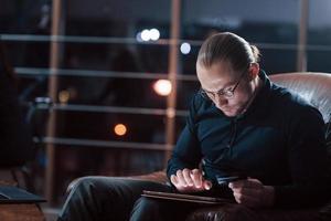 Long haired man. Stylish businessman in eyewear works alone in the office at nightime photo