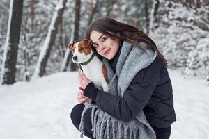 Girl looking to the camera, pet to the side. Smiling brunette having fun while walking with her dog in the winter park photo