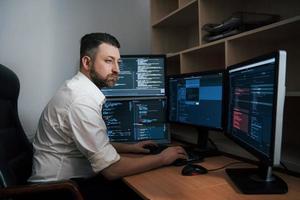 códigos de programa está en todas partes. hombre barbudo con camisa blanca trabaja en la oficina con múltiples pantallas de computadora en gráficos de índice