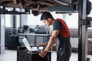 Black hair. Man at the workshop in uniform using laptop for his job for fixing broken car photo