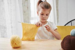 artesanía y pasatiempo para un niño. el niño está tejiendo en casa. linda niña sentada cerca de la mesa de madera está aprendiendo cosas nuevas foto