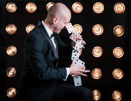 Makes it looks easy. Magician in black suit and with playing cards standing in the room with special lighting at backstage photo