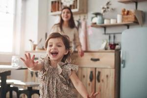 Picture in motion. Playful female child have fun by running in the kitchen at daytime of front of her mother photo