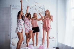 Dancing and laughing. Confetti in the air. Young girls have fun on the white bed in nice room photo