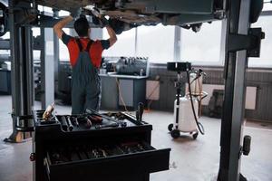 Focused on the table with different instruments. Man at the workshop in uniform fixes broken parts of the modern car photo