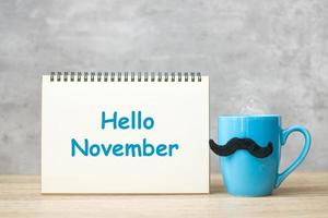 Hello November with paper notepad, Blue coffee cup or tea mug and Black mustache decor on table. International Men day, Happy father day and celebration concept photo