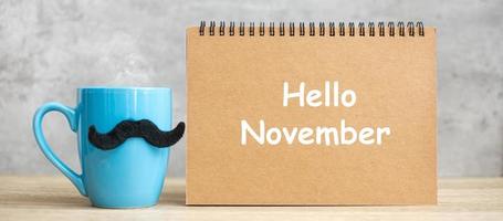 hola noviembre con bloc de notas de papel, taza de café azul o taza de té y decoración de bigote negro en la mesa. día internacional del hombre, feliz día del padre y concepto de celebración foto