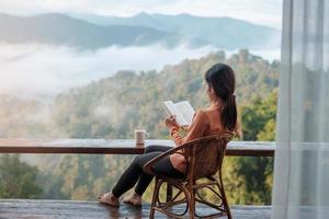 young woman reading book near window and looking mountain view at countryside homestay in the morning sunrise. SoloTravel, journey, trip and relaxing concept photo