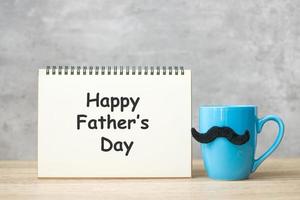 feliz día del padre con bloc de notas de papel, taza de café azul o taza de té y decoración de bigote negro en la mesa. día internacional del hombre y concepto de celebración foto