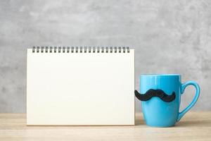 taza de café azul o taza de té con decoración de bigote negro y bloc de notas de papel vacío o calendario en la mesa. espacio de copia en blanco para texto. noviembre azul, feliz día del padre y concepto del día internacional del hombre foto