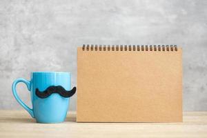 taza de café azul o taza de té con decoración de bigote negro y bloc de notas de papel vacío o calendario en la mesa. espacio de copia en blanco para texto. noviembre azul, feliz día del padre y concepto del día internacional del hombre foto