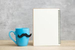 Blue coffee cup or tea mug with Black mustache decor and empty paper notepad or calendar on table. Blank copy space for text. Blue November, Happy Father day and International men day concept photo