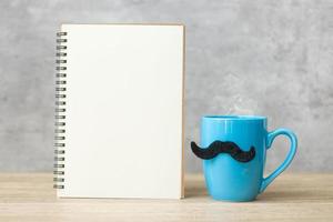 Blue coffee cup or tea mug with Black mustache decor and empty paper notepad or calendar on table. Blank copy space for text. Blue November, Happy Father day and International men day concept photo