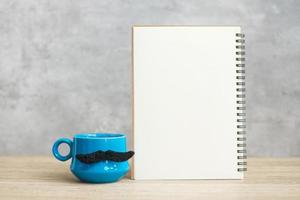 Blue coffee cup or tea mug with Black mustache decor and empty paper notepad or calendar on table. Blank copy space for text. Blue November, Happy Father day and International men day concept