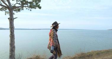 yong hipster kvinna resenär i hatt och poncho promenader utomhus i bergen nära sjön video