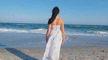 jovem mulher bonita vestida com um vestido branco anda descalço na praia de verão video