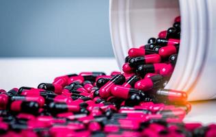 Pink, black capsules pill spilled out from white plastic bottle container. Prescription drug. Antibiotics drug resistance. Antimicrobial capsule pills. Pharmaceutical industry. Pharmacy background. photo