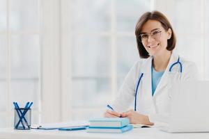 Professional woman doctor writes down notes, poses at desktop in office with laptop, wears white coat, spectacles and phonendoscope around neck, looks through medical documents. Healthcare concept