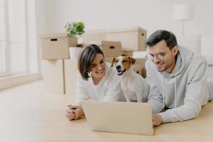 Cheerful husband and wife watch film online on laptop, rest on floor, relax and talk, their domestic animal poses between, relocate in new home, pose in spacious living room with unpacked boxes photo