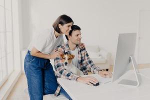 Caring young happy woman leans at husbands shoulders, worries about him, focused into computer monitor, keyboard and surf internet, pose at workplace in modern apartment, little dog sits near photo