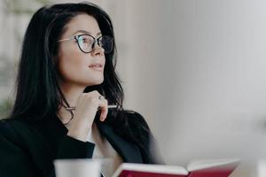 Thoughtful European woman makes notes in notepad, holds pen and writes down some ideas, makes list of goals, plans day in personal organizer, wears transparent glasses, thinks about something photo