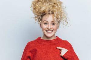 Happy funny beautiful young woman with pleased expression, rejoices her new purchase, indicates at red sweater, shows copy space for your advertisment content. People, style and clothing concept photo