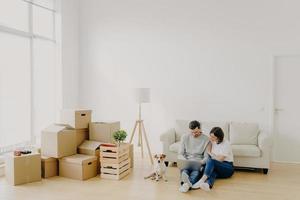 Young relaxed couple move to new home with their favourite pet, sit on floor, surf internet via laptop computer, have day of relocation, surrounded with cardboard boxes filled with personal belongins photo