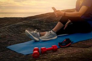 Woman sit and relax on yoga mat near red dumbbells and listen to music via smartphone at stone beach by the sea. Fit girl wear smart band. Healthy, chill out lifestyle. Relax on summer vacations. photo