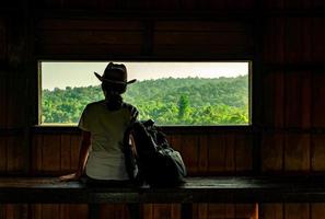 una joven asiática usa el sombrero sentada en un banco de madera y observa una hermosa vista del bosque tropical en la torre de observación de la vida silvestre por la noche con la cálida luz del sol. mujer viaja sola concepto. foto