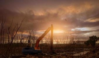 Excavator dredging mud at mangrove. Backhoe digging mud at construction site. Excavating machine removing sediment from waterway. Excavation vehicle. Backhoe working in evening with sunset sky. photo