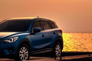 Blue compact SUV car with sport and modern design parked on concrete road by sea at sunset. Environmentally friendly technology. Hybrid and electric car technology. Car parking space. summer travel. photo