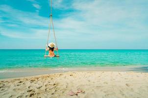 Asian women wear swimwear and hat swing the swings at sand beach and looking beautiful tropical paradise sea and sky on a sunny day. Summer vacation. Summer vibes. Enjoying and relaxing girl photo