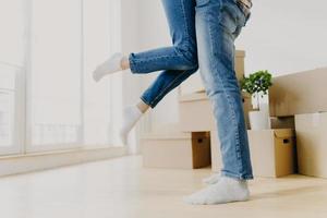 Happy unknown couple move in new abode, man lifts woman, wear jeans, pose in empty room with carton boxes around, start living seperate from parents rejoice buying apartment. Unrecognizable homeowners photo