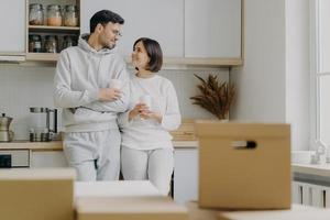 Image of cheerful woman and man have pleasant talk during coffee break, wear casual clothes, move in new apartment, pose with modern furnished kitchen, unpack cardboard boxes with household stuff photo