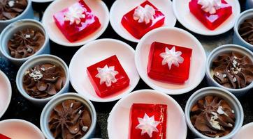 Red jelly dessert with whipped cream served on white plate and cupcakes with chocolate mousse in cup. Square red jelly plate on table at restaurant for buffet. Sweet food. Jelly agar, selective focus. photo