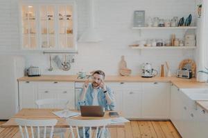 Young office worker working remotely from home with computer while sitting by kitchen table photo