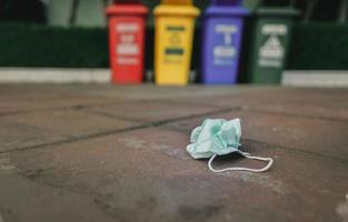 Used medical face mask discard on pavement floor on blurred recycle bin or trash. Medical waste disposal with unhygienic. Disposable earloop face mask waste garbage on floor. Green surgical face mask. photo