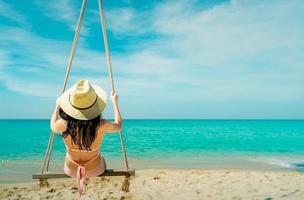 Asian women wear swimwear and hat swing the swings at sand beach and looking beautiful tropical paradise sea and sky on a sunny day. Summer vacation. Summer vibes. Enjoying and relaxing girl photo