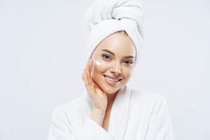Charming relaxed beauty woman applies face cream, cares about complexion, touches cheek with hand, smiles gently at camera, dressed in bath robe, wrapped towel on washed hair, isolated on white photo