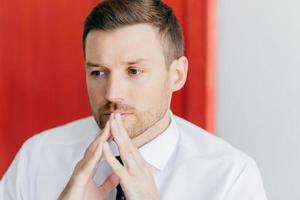 Thoughtful attractive male keeps hands together near mouth, has pensive expression, dressed in white shirt, thinks about something, isolated over red and white background. People and career concept