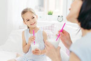 Cropped shot of beautiful small female child drinks tasty milk shake together with her mother, enjoys good morning and calm domestic atmosphere, has happy look. People, breakfast and bedding photo