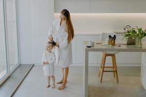 madre cariñosa peina el cabello de su hija pequeña usa albornoces blancos posan contra el interior de la cocina descalza en el suelo. hermosa niña rizada después de tomar una ducha. mamá haciendo peinado a niño foto