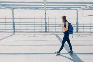 Horizontal shot of good looking sportswoman dressed in casual outfit, carries rucksack, being addicted to modern technologies, always in touch, listens favourite song. Active lifestyle concept photo