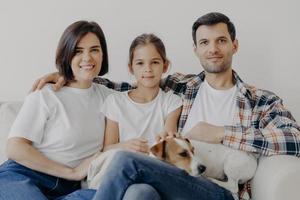 Portrait of affectionate family cuddle and sit together at couch in living room, change their home, have happy expressions. Father, mother, daughter and dog pose for making portrait, spend good time photo