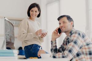 Young married couple study contract for new house, check documents for buying property, man discusses something with partner via cellphone, happy woman drinks tea and looks in computer monitor photo