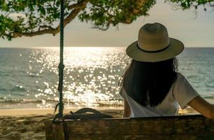 Young Asian woman sit and relax on swings at seaside on summer vacation. Summer vibes. Woman travel alone on holiday. Backpacker enjoy life at tropical paradise beach. Back view of woman watch sunset photo