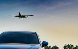 Commercial airline. Passenger plane landing approach blue SUV car at airport with blue sky and clouds at sunset. Arrival flight. Vacation time. Happy trip. Airplane flying on bright sky. Car parked. photo