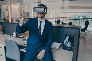 Businessman sitting on desk in office and using VR headset on his head, pointing with forefinger photo
