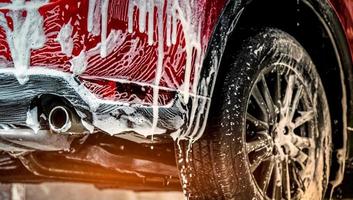 Red compact SUV car with sport and modern design washing with soap. Car covered with white foam. Car care service business concept. Car wash with foam before glass waxing and glass coating automobile. photo